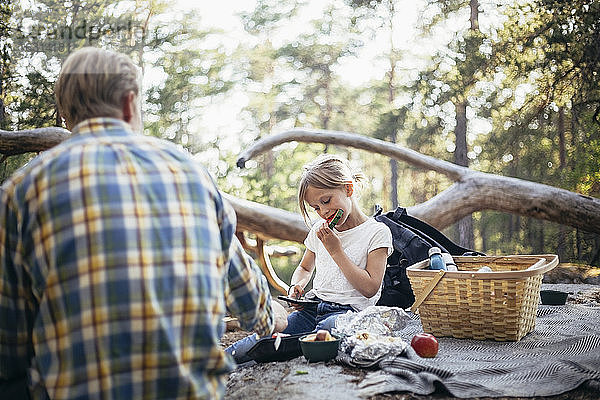 Tochter isst Wassermelone  während sie mit dem Vater auf einer Picknickdecke im Wald sitzt