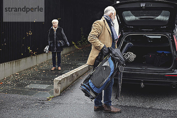 Älterer Mann hält Golftasche am Kofferraum des Autos  während die Partnerin im Winter auf dem Bürgersteig steht