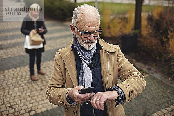 Älterer Mann mit Smartphone  der die Zeit überprüft  während sein Partner auf dem Fußweg hinter ihm steht