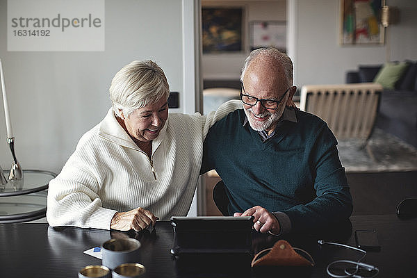 Lächelndes älteres Ehepaar benutzt digitales Tablett  während es am Esstisch im Wohnzimmer sitzt