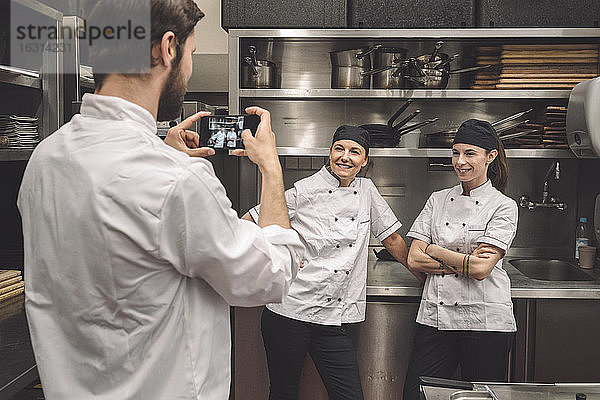 Männlicher Koch fotografiert lächelnde weibliche Mitarbeiter in einer Großküche