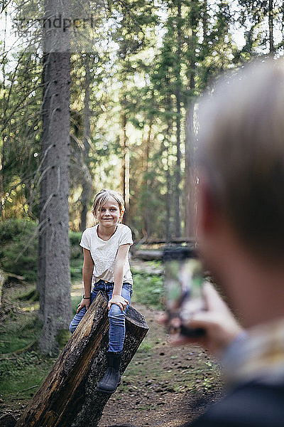 Lächelnde Tochter sitzt auf einem Baumstamm  während der Vater im Wald mit einem Smartphone fotografiert