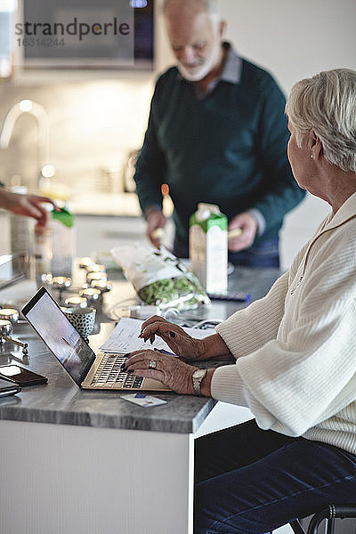 Ältere Frau benutzt Laptop  während der männliche Partner an der Kücheninsel steht