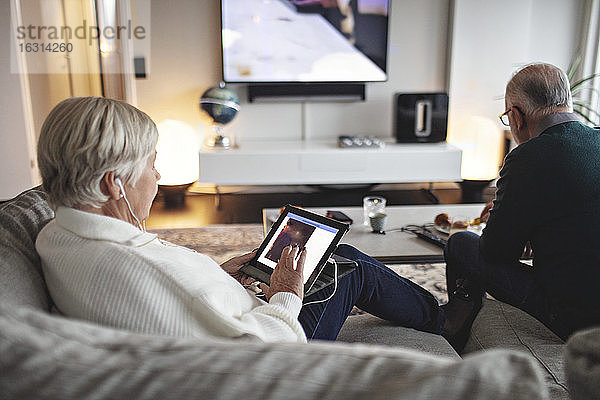 Ältere Frau benutzt digitales Tablett  während der männliche Partner auf dem Sofa im Wohnzimmer sitzt