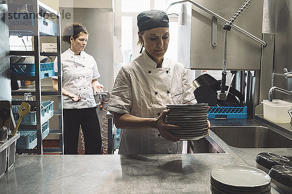 Köchin hält Teller  während eine Mitarbeiterin in der Großküche im Hintergrund steht