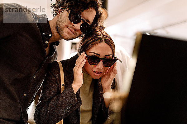 Lächelnde Frau probiert mit Freundin im Geschäft eine modische Sonnenbrille aus