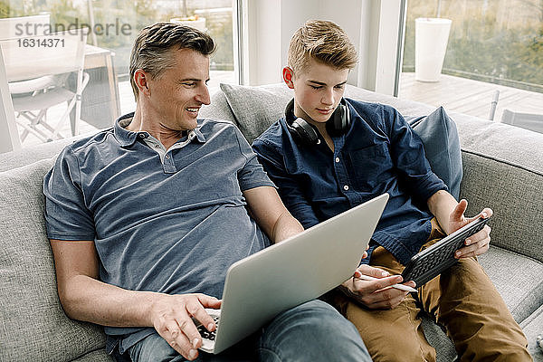 Lächelnder Vater mit Laptop  der den Sohn mit einem digitalen Tablet anschaut  während er zu Hause auf dem Sofa sitzt