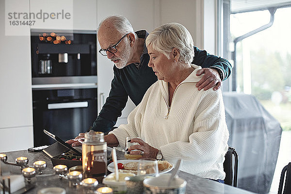 Älteres Ehepaar betrachtet digitales Tablett  während es am Esstisch im Wohnzimmer sitzt