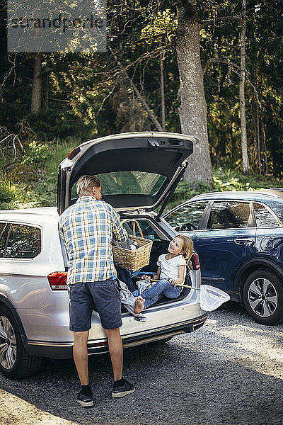 Rückansicht des Vaters stehend mit Picknickkorb  während die Tochter im Kofferraum des Autos sitzt