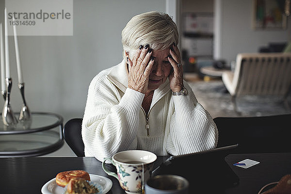Besorgte ältere Frau mit dem Kopf in der Hand  die auf ein digitales Tablet schaut  während sie zu Hause am Esstisch sitzt