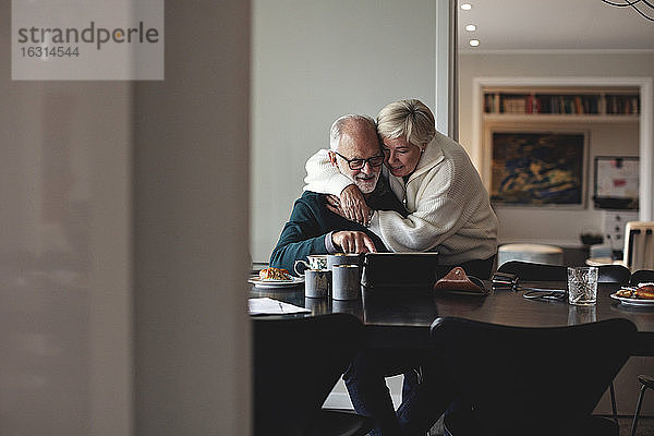 Lächelnde ältere Frau umarmt Partner am Esstisch im Wohnzimmer