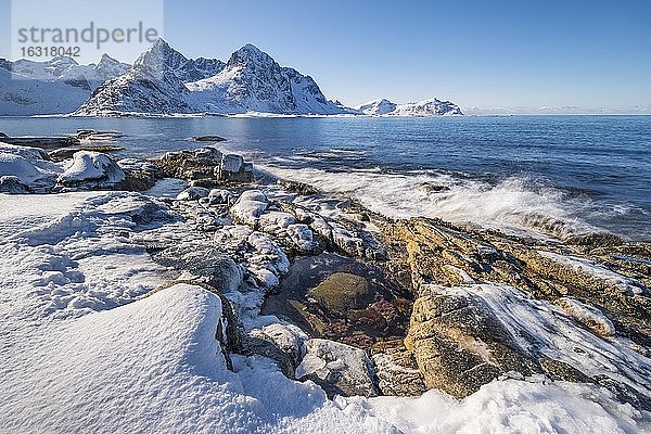 Verschneiter Felsstrand mit Gezeitentümpel  Küste mit blauem Meer  hinten winterliche Berge  Nordland  Lofoten  Norwegen  Europa