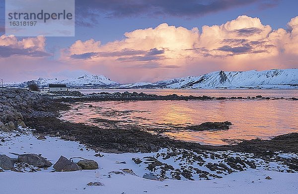 Verschneite Landschaft mit Holzhaus an Meeresbucht und Fjord  Wolkenstimmung  warmes Abendlicht  Nordland  Lofoten  Norwegen  Europa