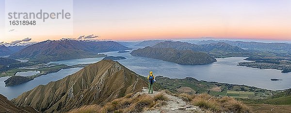 Junger Mann steht auf dem Gipfel  Ausblick auf Berge und See vom Mount Roy  Roys Peak bei Sonnenuntergang  Lake Wanaka  Südalpen  Otago  Südinsel  Neuseeland  Ozeanien