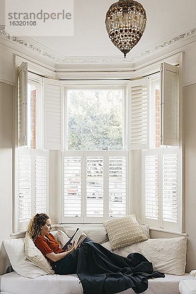Junge Frau entspannt sich auf dem Wohnzimmersofa und liest ein Buch im Erkerfenster