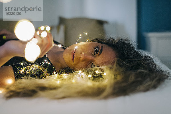 Porträt heitere junge Frau mit Lichterkette auf Bett