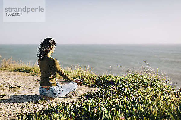 Gelassene Frau meditiert auf einer sonnigen Klippe mit Blick auf das Meer