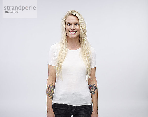 Porträt einer lächelnden blonden jungen Frau mit Tätowierung an den Unterarmen vor einem hellen Hintergrund
