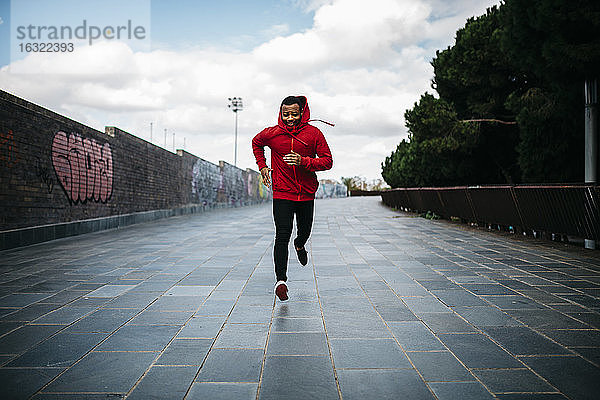 Lächelnder junger Mann mit rotem Kapuzenpulli beim Laufen in der Stadt