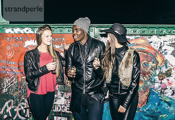 Freunde mit Sektgläsern an einer nächtlichen Graffiti-Wand stehend