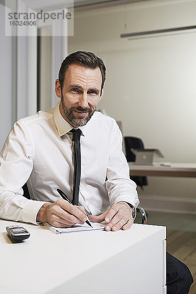 Porträt eines selbstbewussten Geschäftsmannes  der im Büro am Schreibtisch sitzt und Notizen macht