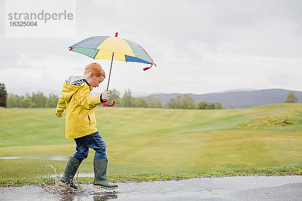 Rothaariger kleiner Junge mit Regenschirm spielt im Regen