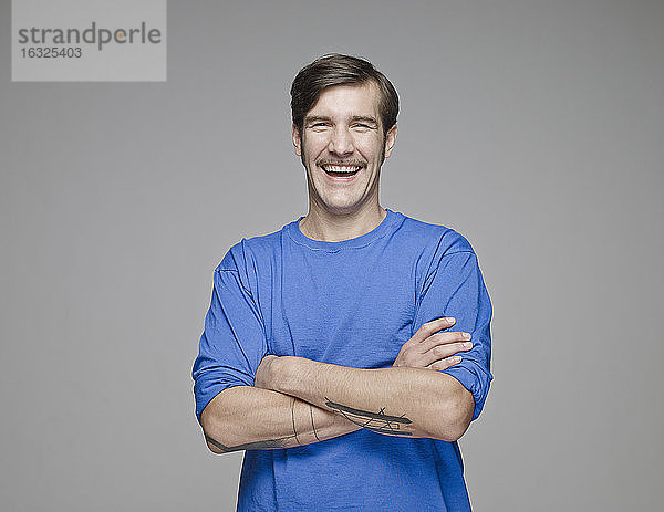 Porträt eines lachenden Mannes mit verschränkten Armen vor einem grauen Hintergrund
