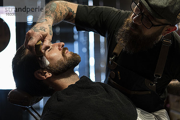 Friseur trägt Rasierschaum auf den Bart eines Kunden auf
