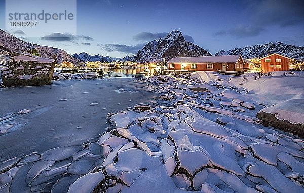 Eisschollen am zugefrorenem Fjord  hinten rote Holzhäuser und winterliche Berglandschaft in der Abenddämmerung  Reine  Nordland  Lofoten  Norwegen  Europa
