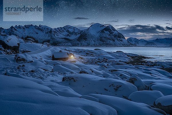 Tief verschneite Winterlandschaft am Meer  Holzhütte mit warmem Licht an Küste  hinten Berge unter Sternenhimmel  Kleppstad  Nordland  Lofoten  Norwegen  Europa