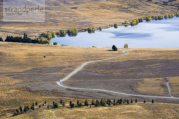 Foto eines Road-Trips in einem Wohnwagen am Lake Alexandrina auf der Südinsel  Neuseeland. Dieses Foto eines Wohnwagens  der sich dem Alexandrina-See auf einer langen verlassenen Straße nähert  wurde vom St. John Observatory auf der Südinsel Neuseelands aufgenommen. Vom St. John Observatory aus hat man einen unvergleichlichen Blick auf den Tekapo-See  den Alexandrina-See und die umliegende Landschaft mit ihren schneebedeckten Bergen und Baron-Feldern  die entstanden  als sich die Seen durch den Rückzug der Gletscher bildeten. Neuseeland  und insbesondere die Südinsel Neuseelands  ist eine fantastische Kulisse für einen Roadtrip im Wohnwagen oder Wohnmobil. Morgens wachen Sie mit einem atemberaubenden Blick auf ruhige  neblige Seen auf und müssen kaum aus dem Wohnmobil steigen  um die unglaublichen Landschaften zu erleben.