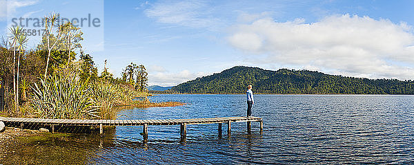 Panoramafoto eines Touristen  der die Ruhe am Lake Ianthe genießt  Westküste  Südinsel  Neuseeland. Dieses Panoramafoto wurde am Lake Ianthe aufgenommen  einem der zahlreichen Höhepunkte der Fahrt entlang der Westküste der neuseeländischen Südinsel.