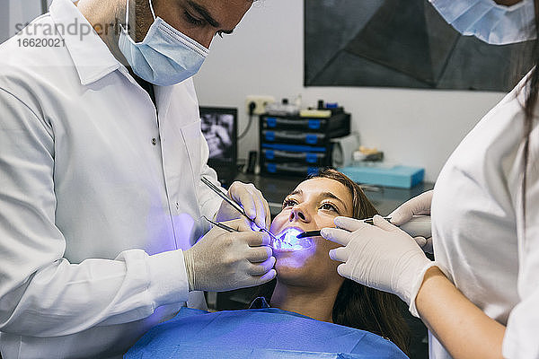 Männlicher Zahnarzt in Maske und Handschuhen untersucht die Zähne einer Patientin mit Hilfe einer Assistentin in einer Klinik