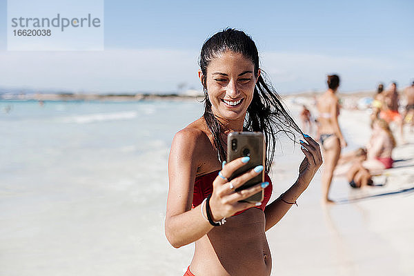 Lächelnde Frau im Bikini macht Selfie mit Handy am Strand gegen den Himmel