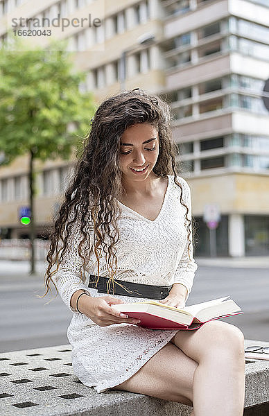 Schöne Frau mit langen Haaren  die ein Buch liest  während sie auf einem Sitz in der Stadt sitzt