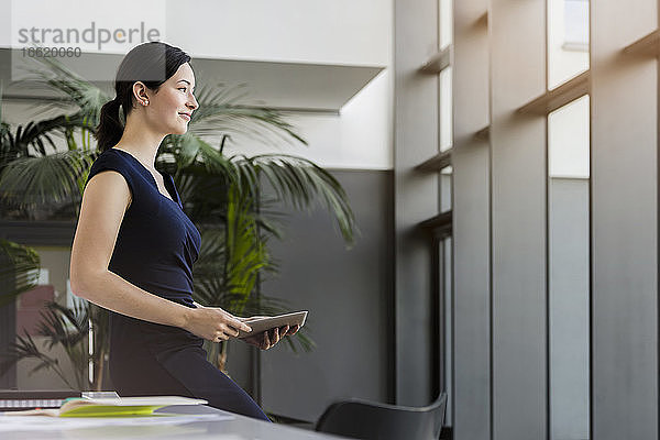 Nachdenkliche Geschäftsfrau  die ein digitales Tablet hält  während sie auf einem Schreibtisch im Büro sitzt