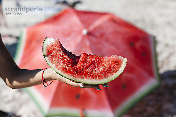 Hand einer Frau  die eine Wassermelone gegen einen roten Regenschirm am Strand hält