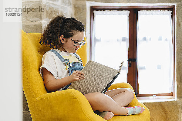 Mädchen liest ein Buch  während sie zu Hause auf einem Sessel sitzt