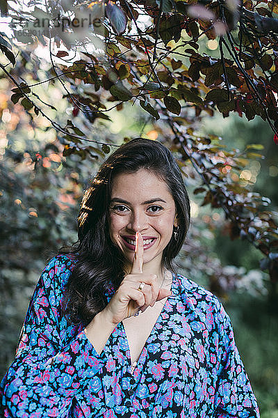 Nahaufnahme einer lächelnden jungen Frau mit Finger auf den Lippen  die im Park vor Pflanzen steht