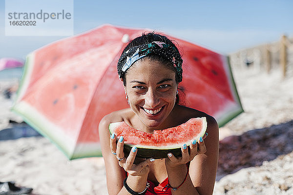 Nahaufnahme einer lächelnden Frau  die eine Wassermelone in der Hand hält  während sie unter einem roten Sonnenschirm am Strand sitzt