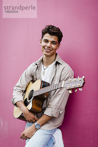 Glücklicher Mann hält Gitarre und lehnt sich an eine rosa Wand