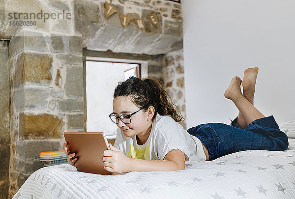 Mädchen liegt auf der Vorderseite und benutzt ein digitales Tablet zu Hause im Schlafzimmer