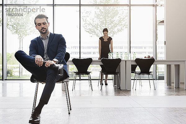 Seriöser Geschäftsmann sitzt auf einem Stuhl  während eine Mitarbeiterin im Hintergrund am Schreibtisch arbeitet