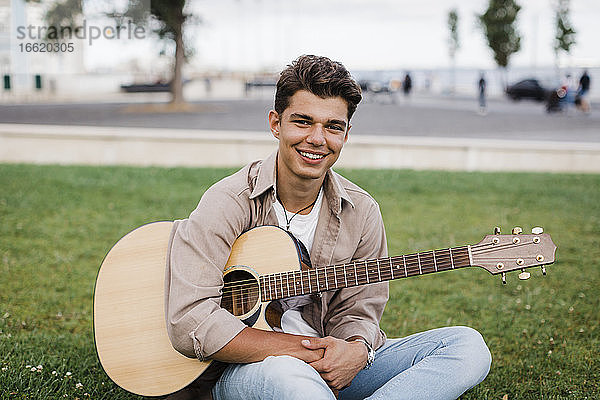 Lächelnder gut aussehender junger Mann sitzt mit Gitarre im Gras im Park
