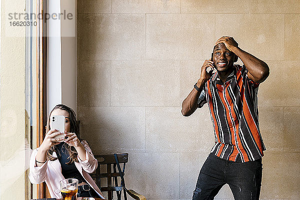 Junger Mann mit Hand auf dem Kopf  während er mit seiner Freundin im Café über sein Smartphone spricht
