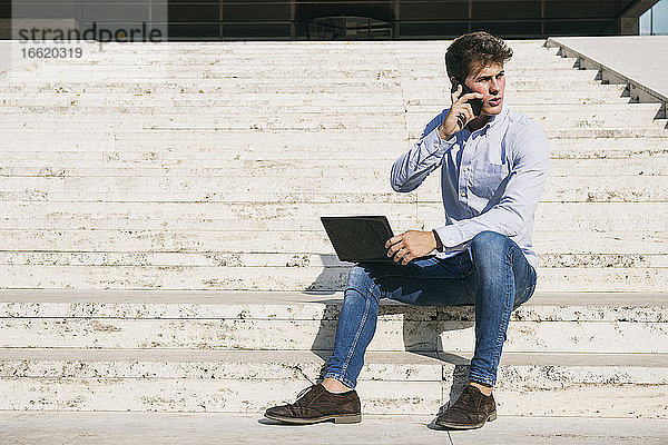 Gut aussehender junger Mann  der mit seinem Laptop auf einer Treppe sitzt und über sein Handy spricht