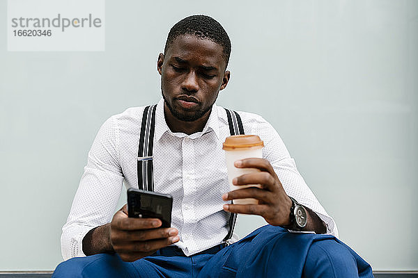 Junger Geschäftsmann  der ein Smartphone benutzt  während er einen wiederverwendbaren Becher in der Hand hält und gegen ein Fenster in der Stadt sitzt