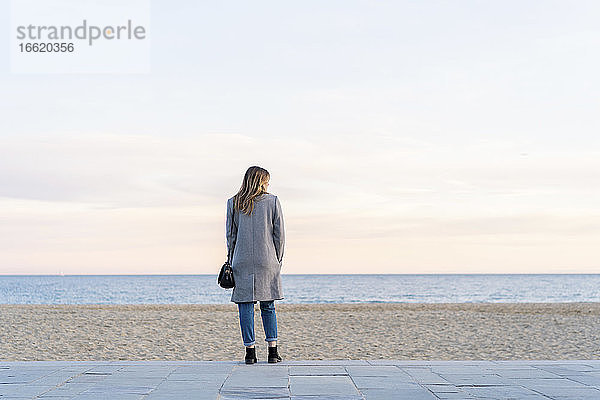 Junge Frau schaut auf das Meer  während sie auf der Promenade am Strand steht und den Sonnenuntergang genießt