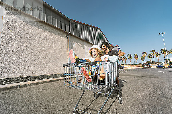 Junger Mann schiebt seine Freundin im Einkaufswagen vor einem Supermarkt