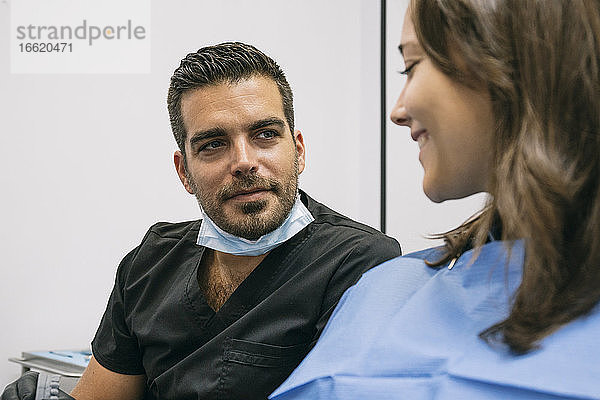 Gut aussehender männlicher Zahnarzt erklärt einer lächelnden Patientin in der Praxis die Zahnbehandlung